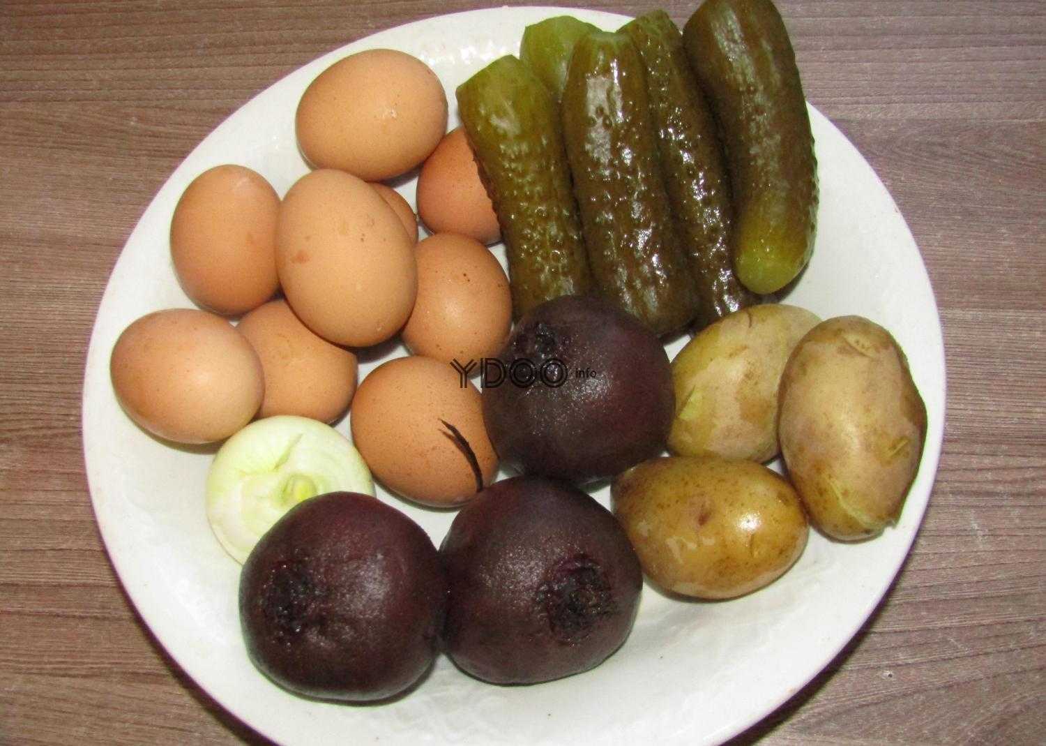 три вареных свеклы, три вареных картофелины, один репчатый лук, яйца куриные, соленые огурцы на белой круглой тарелке