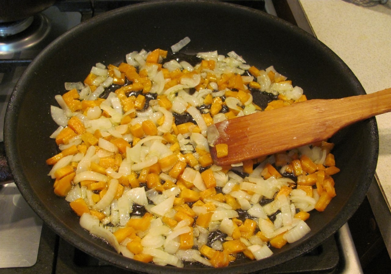жареные лук с морковью в сковороде с маслом и деревянной лопаткой