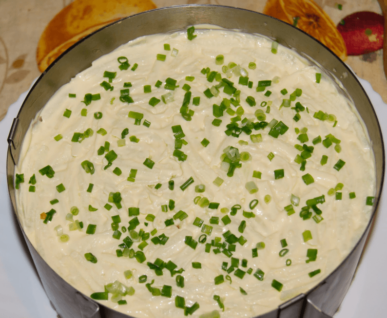 на большой тарелке лежит кулинарной кольцо, внутри слоеный салат селедка под шубой, сверху блюдо присыпано измельченным зеленым луком